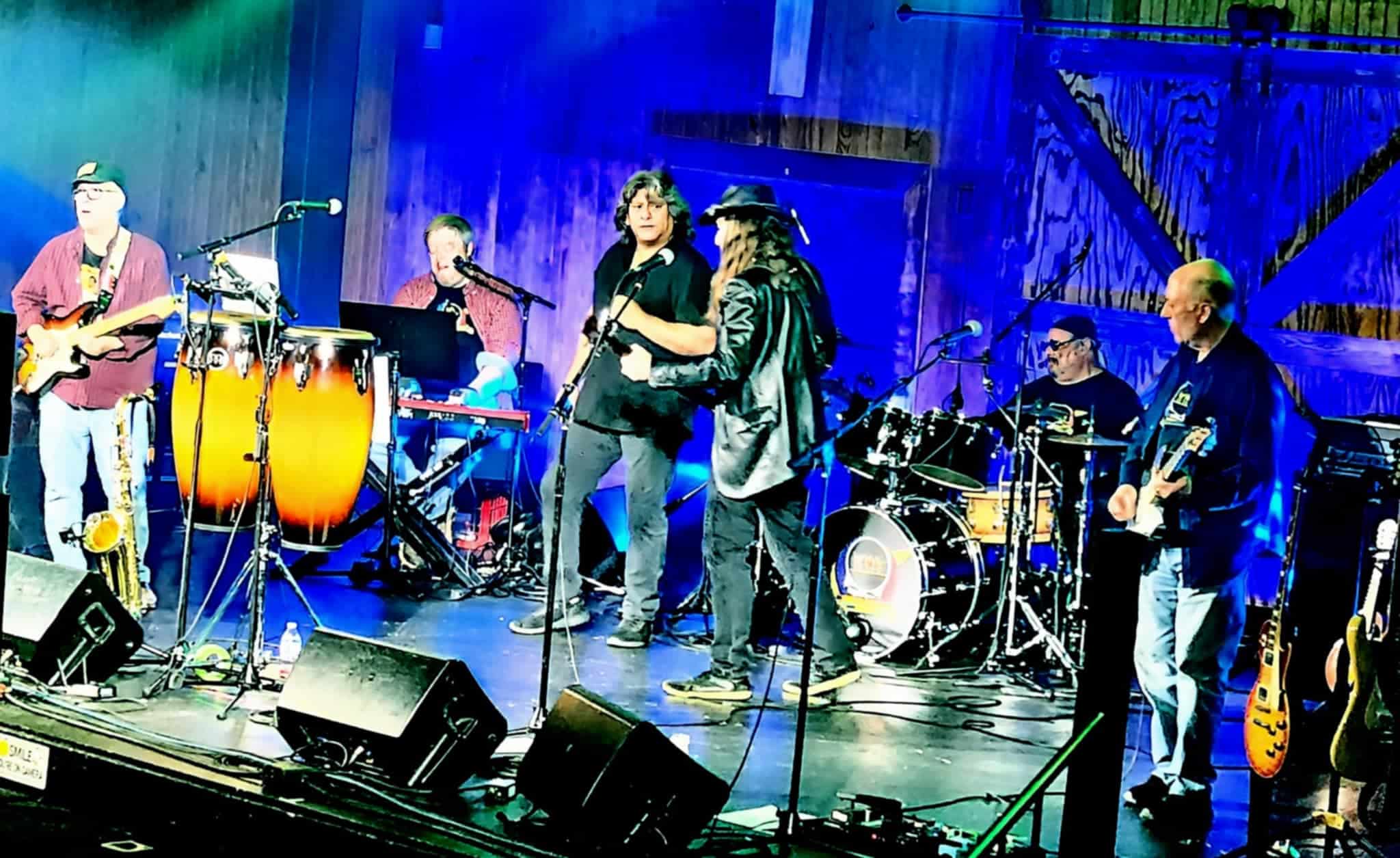 A lively band performs on stage with guitars, drums, and congas. Bright stage lights highlight their energetic vibe amid a wooden backdrop.